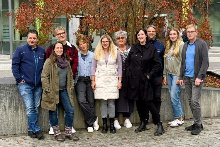 Eine fröhliche Gruppe steht vor herbstlich rot eingefärbten Baum und lächelt in die Kamera. Im Hintergrund ist die Bibliothek des Campus Kamp-Lintfort mit den grünen Fensterrahmen zu erkennen.