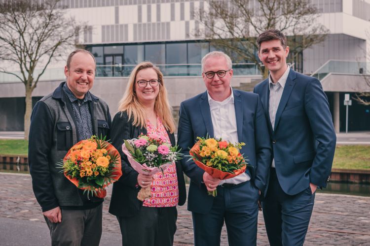 Vier Personen stehen nebeneinander vor dem Hörsaalzentrum der Hochschule Rhein-Waal und lächeln in die Kamera. Drei von ihnen halten einen Blumenstrauß.