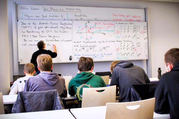 Nachwuchsmathetalente schauen in der Mathematikvorlesung auf die Tafel, wo eine wissenschaftliche Lehrkraft mathematische Gleichungen anschreibt.