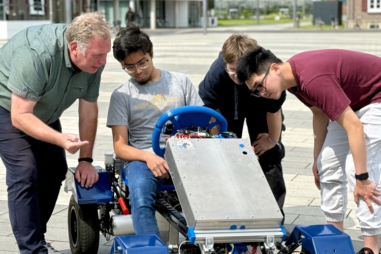 Studiengangsleiter Prof. Dr. Nissing testet zusammen mit drei Studierenden ein E-Gokart. Ein Studierender sitzt auf dem GoKart und schaut auf die Pedale. Zwei weitere Studierende beugen sich zum Lenkrad herunter. Professor Nissing erklärt vorgebeugt die Funktionen.