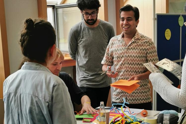 Teilnehmende der StartGlocal Summerschool beim gemeinsamen Brainstorming. Fünf Personen stehen um einen Tisch mit buntem Papier und unterhalten sich angeregt.