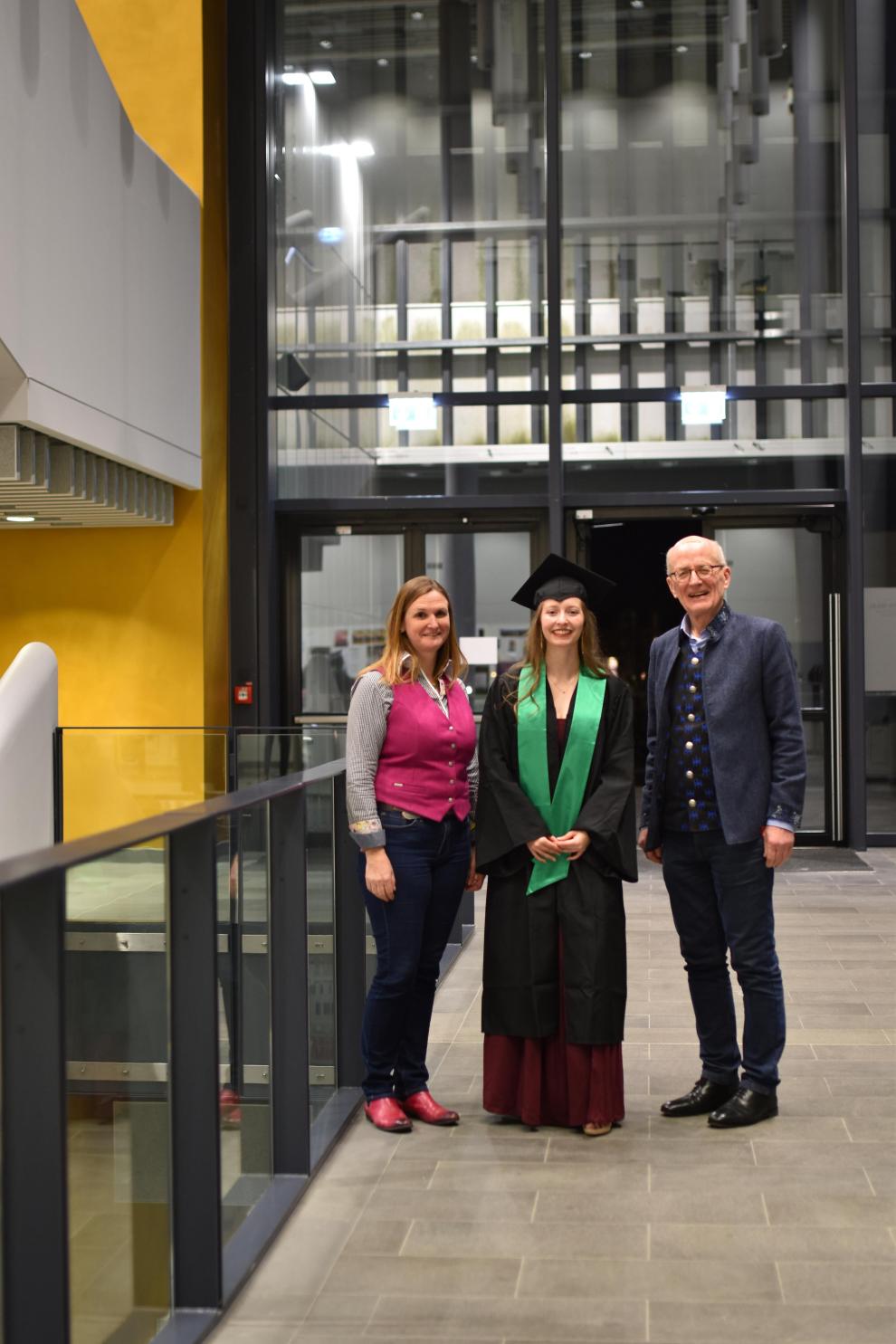Prof. Simone Pauling, die Preisträgerin in Robe und Prof. Schuhmacher stehen nebeneinander im ersten Stock des Hochschulgebäudes